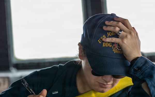 A U.S. Navy sailor in dark blue coveralls is seated at a desk aboard a naval vessel, writing in or reviewing a binder labeled 'NAV' (Navigation). The sailor is wearing a cap bearing 'USS John...' and 'DDG' designation, along with sunglasses and a yellow inflatable life vest/collar. The setting appears to be the bridge or a navigation workspace of a Navy destroyer.