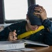 A U.S. Navy sailor in dark blue coveralls is seated at a desk aboard a naval vessel, writing in or reviewing a binder labeled 'NAV' (Navigation). The sailor is wearing a cap bearing 'USS John...' and 'DDG' designation, along with sunglasses and a yellow inflatable life vest/collar. The setting appears to be the bridge or a navigation workspace of a Navy destroyer.