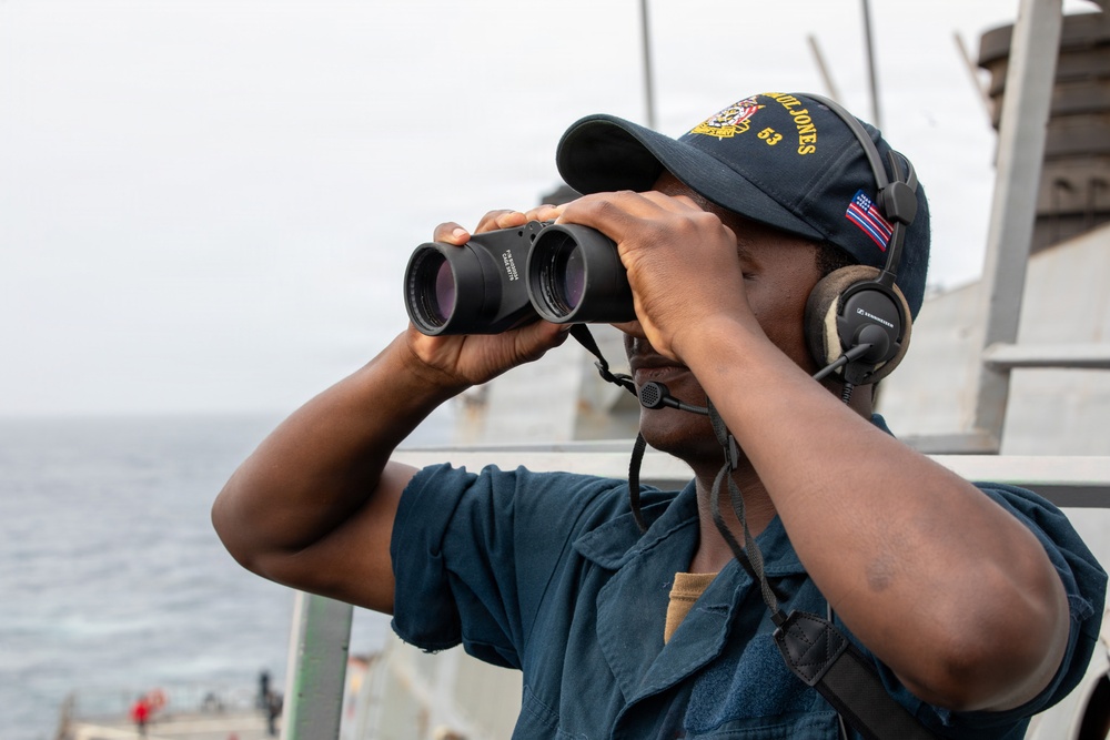 A U.S. Navy sailor is standing on the deck of a naval vessel, using binoculars to scan the horizon over open water. The sailor is wearing a navy blue uniform, a ship's ball cap with the number '63' visible, and Sennheiser communications headphones. The image appears to be an official military photograph taken aboard a U.S. Navy destroyer or similar warship.