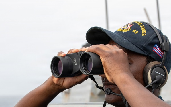 A U.S. Navy sailor is standing on the deck of a naval vessel, using binoculars to scan the horizon over open water. The sailor is wearing a navy blue uniform, a ship's ball cap with the number '63' visible, and Sennheiser communications headphones. The image appears to be an official military photograph taken aboard a U.S. Navy destroyer or similar warship.