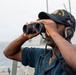 A U.S. Navy sailor is standing on the deck of a naval vessel, using binoculars to scan the horizon over open water. The sailor is wearing a navy blue uniform, a ship's ball cap with the number '63' visible, and Sennheiser communications headphones. The image appears to be an official military photograph taken aboard a U.S. Navy destroyer or similar warship.