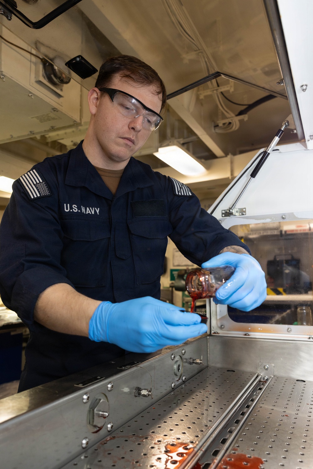 A U.S. Navy sailor wearing a dark blue uniform, safety glasses, and blue nitrile gloves is pouring a reddish-brown liquid from a small glass container into a metal equipment housing. The work appears to be taking place in a shipboard maintenance or technical workspace. The activity suggests a fluid sampling, lubrication, or maintenance procedure on naval equipment.