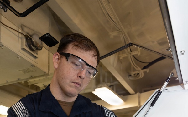 A U.S. Navy sailor wearing a dark blue uniform, safety glasses, and blue nitrile gloves is pouring a reddish-brown liquid from a small glass container into a metal equipment housing. The work appears to be taking place in a shipboard maintenance or technical workspace. The activity suggests a fluid sampling, lubrication, or maintenance procedure on naval equipment.