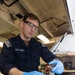 A U.S. Navy sailor wearing a dark blue uniform, safety glasses, and blue nitrile gloves is pouring a reddish-brown liquid from a small glass container into a metal equipment housing. The work appears to be taking place in a shipboard maintenance or technical workspace. The activity suggests a fluid sampling, lubrication, or maintenance procedure on naval equipment.
