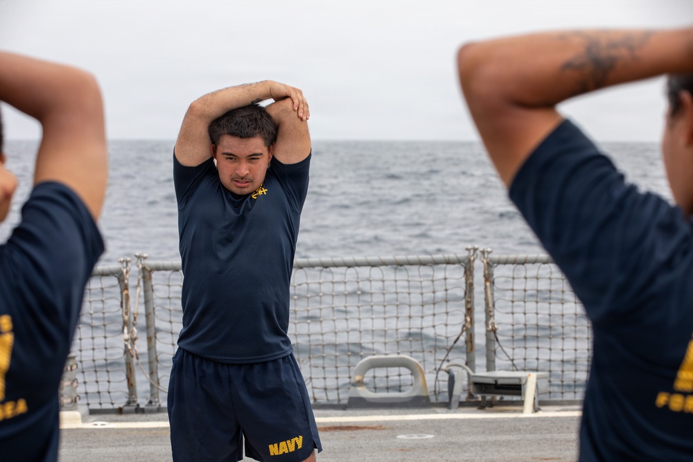 Several U.S. Navy personnel are performing stretching exercises on the deck of a naval vessel at sea. The individuals are wearing standard Navy physical training (PT) uniforms consisting of dark navy blue shirts and shorts with 'NAVY' printed in gold lettering. The overcast sky and open ocean are visible in the background beyond the ship's safety railing.