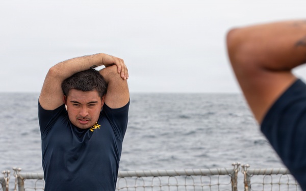 Several U.S. Navy personnel are performing stretching exercises on the deck of a naval vessel at sea. The individuals are wearing standard Navy physical training (PT) uniforms consisting of dark navy blue shirts and shorts with 'NAVY' printed in gold lettering. The overcast sky and open ocean are visible in the background beyond the ship's safety railing.