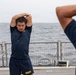 Several U.S. Navy personnel are performing stretching exercises on the deck of a naval vessel at sea. The individuals are wearing standard Navy physical training (PT) uniforms consisting of dark navy blue shirts and shorts with 'NAVY' printed in gold lettering. The overcast sky and open ocean are visible in the background beyond the ship's safety railing.