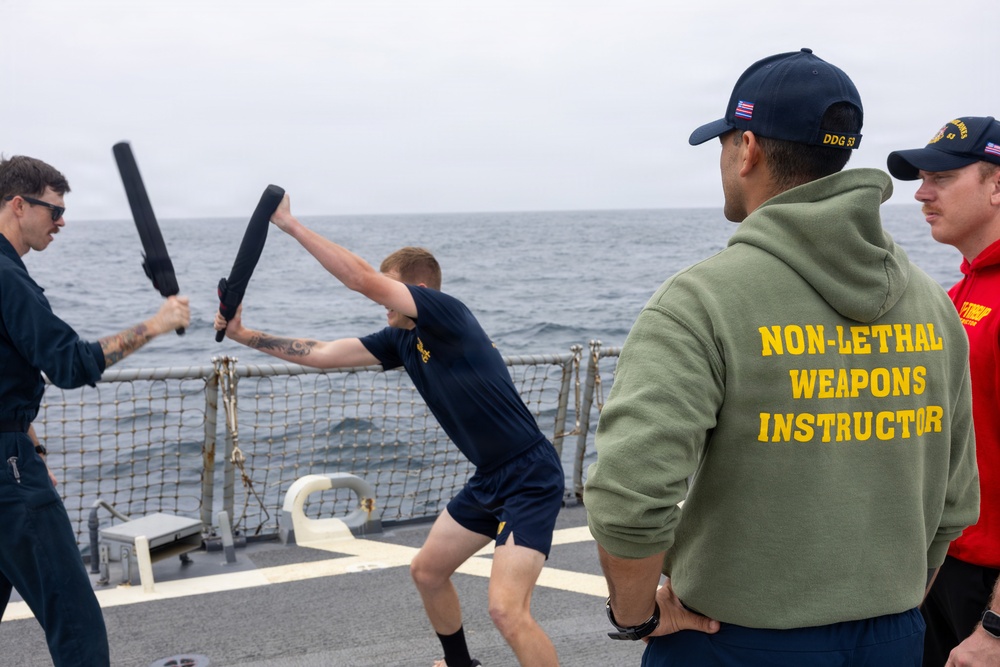 A non-lethal weapons training exercise is taking place on the deck of a U.S. Navy destroyer at sea. Two personnel are sparring with foam/padded batons while an instructor wearing a green hoodie labeled 'NON-LETHAL WEAPONS INSTRUCTOR' observes alongside another crew member. The ship's hull number visible on the cap reads 'DDG 53'.