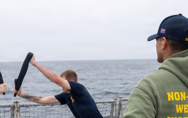 A non-lethal weapons training exercise is taking place on the deck of a U.S. Navy destroyer at sea. Two personnel are sparring with foam/padded batons while an instructor wearing a green hoodie labeled 'NON-LETHAL WEAPONS INSTRUCTOR' observes alongside another crew member. The ship's hull number visible on the cap reads 'DDG 53'.
