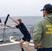 A non-lethal weapons training exercise is taking place on the deck of a U.S. Navy destroyer at sea. Two personnel are sparring with foam/padded batons while an instructor wearing a green hoodie labeled 'NON-LETHAL WEAPONS INSTRUCTOR' observes alongside another crew member. The ship's hull number visible on the cap reads 'DDG 53'.