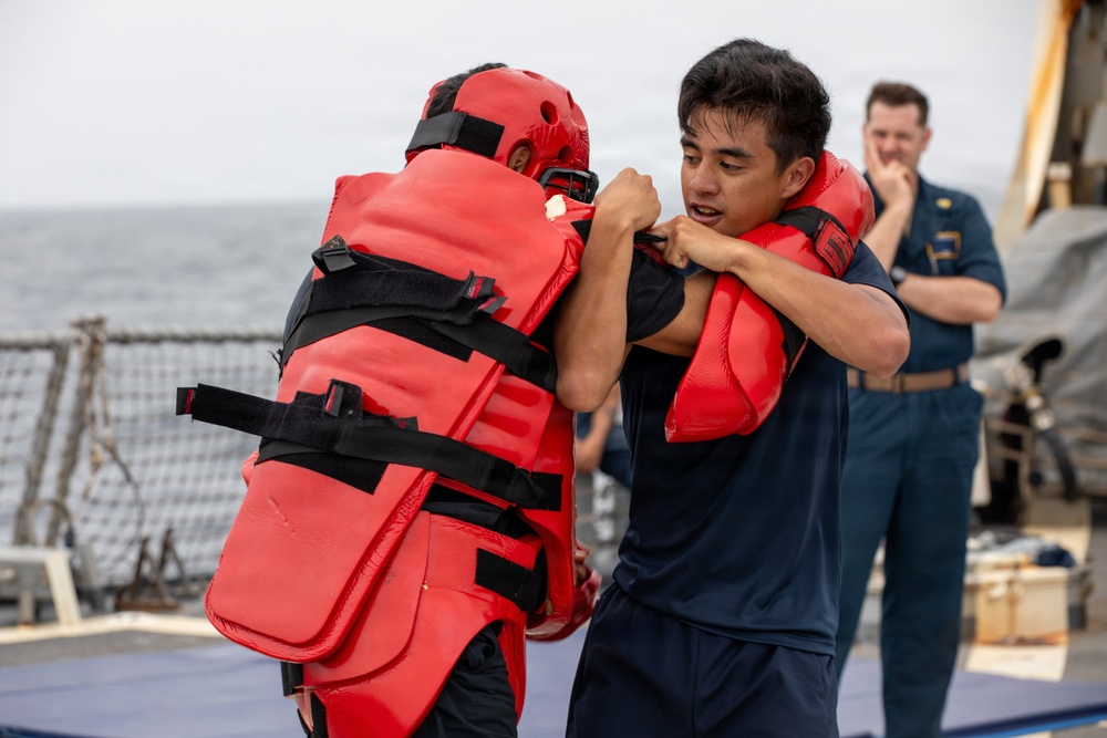 Two individuals are engaged in a close-quarters combat or self-defense training drill on what appears to be the deck of a naval vessel. One participant is wearing full red protective padding and a red helmet, while the other is in a navy blue t-shirt and shorts. A third person in a military uniform observes the drill in the background.