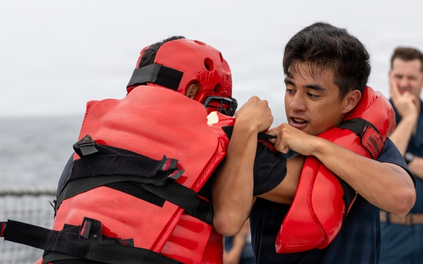 Two individuals are engaged in a close-quarters combat or self-defense training drill on what appears to be the deck of a naval vessel. One participant is wearing full red protective padding and a red helmet, while the other is in a navy blue t-shirt and shorts. A third person in a military uniform observes the drill in the background.