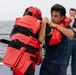 Two individuals are engaged in a close-quarters combat or self-defense training drill on what appears to be the deck of a naval vessel. One participant is wearing full red protective padding and a red helmet, while the other is in a navy blue t-shirt and shorts. A third person in a military uniform observes the drill in the background.