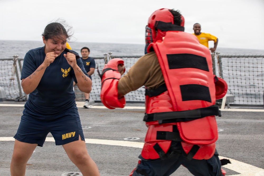 A U.S. Navy sailor in PT (physical training) uniform is practicing combat or self-defense drills on what appears to be the deck of a naval vessel. She is in a fighting stance facing an opponent wearing full red protective padding and a helmet. Several other sailors are visible in the background observing the training exercise.