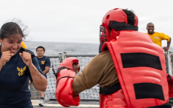 A U.S. Navy sailor in PT (physical training) uniform is practicing combat or self-defense drills on what appears to be the deck of a naval vessel. She is in a fighting stance facing an opponent wearing full red protective padding and a helmet. Several other sailors are visible in the background observing the training exercise.