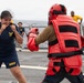 A U.S. Navy sailor in PT (physical training) uniform is practicing combat or self-defense drills on what appears to be the deck of a naval vessel. She is in a fighting stance facing an opponent wearing full red protective padding and a helmet. Several other sailors are visible in the background observing the training exercise.