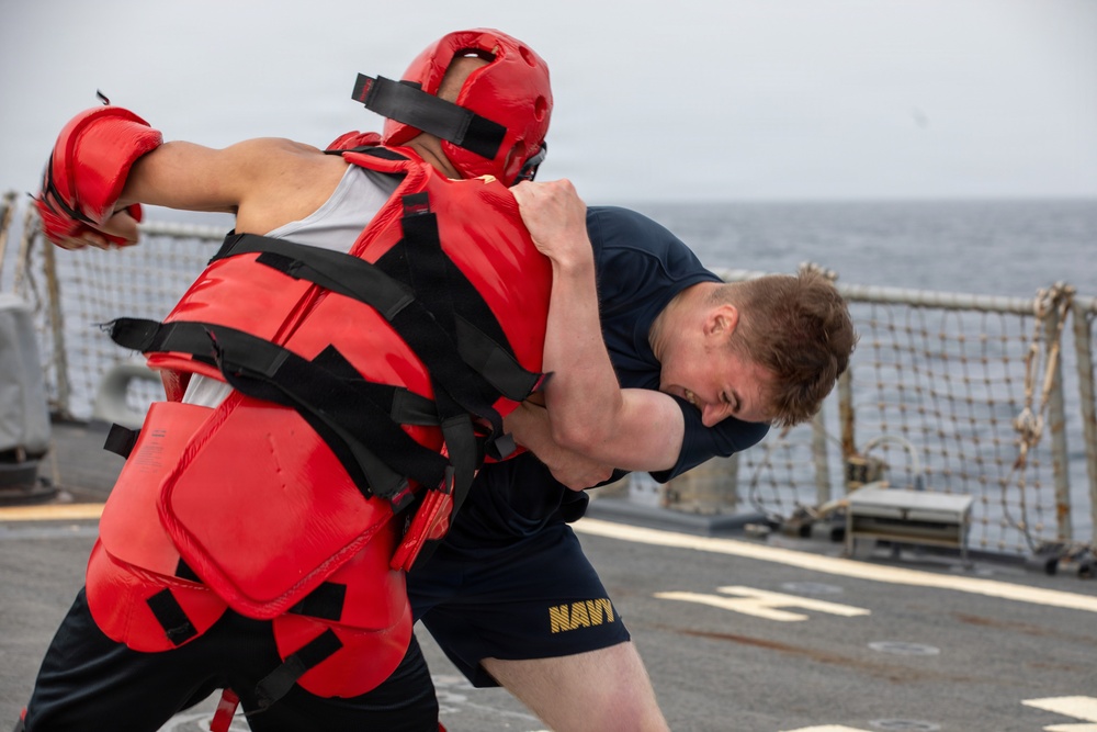 A U.S. Navy sailor in dark blue shorts and t-shirt is engaged in a close-quarters combat training drill on the deck of a naval vessel. His training partner is wearing full red protective gear including a padded helmet, chest protector, and gloves. The drill appears to be taking place at sea, with ocean water visible in the background.