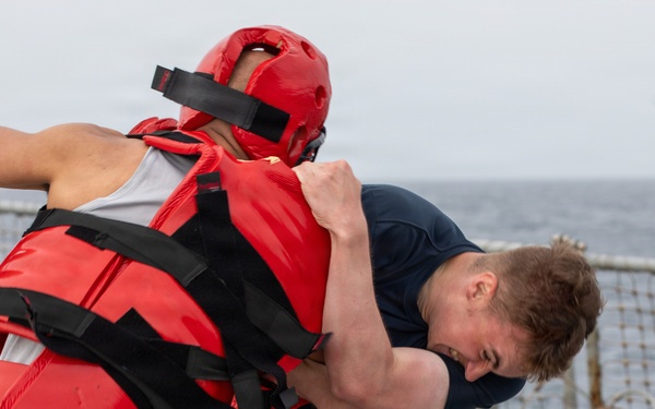 A U.S. Navy sailor in dark blue shorts and t-shirt is engaged in a close-quarters combat training drill on the deck of a naval vessel. His training partner is wearing full red protective gear including a padded helmet, chest protector, and gloves. The drill appears to be taking place at sea, with ocean water visible in the background.