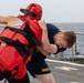 A U.S. Navy sailor in dark blue shorts and t-shirt is engaged in a close-quarters combat training drill on the deck of a naval vessel. His training partner is wearing full red protective gear including a padded helmet, chest protector, and gloves. The drill appears to be taking place at sea, with ocean water visible in the background.