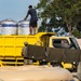 A yellow truck carries large cylindrical water storage tanks, with a civilian man standing on top of the truck bed managing the tanks. Military personnel in camouflage uniforms are on the ground handling hoses, suggesting a water distribution or filling operation. The scene appears to be a field logistics or humanitarian aid operation, likely in a Southeast Asian setting.