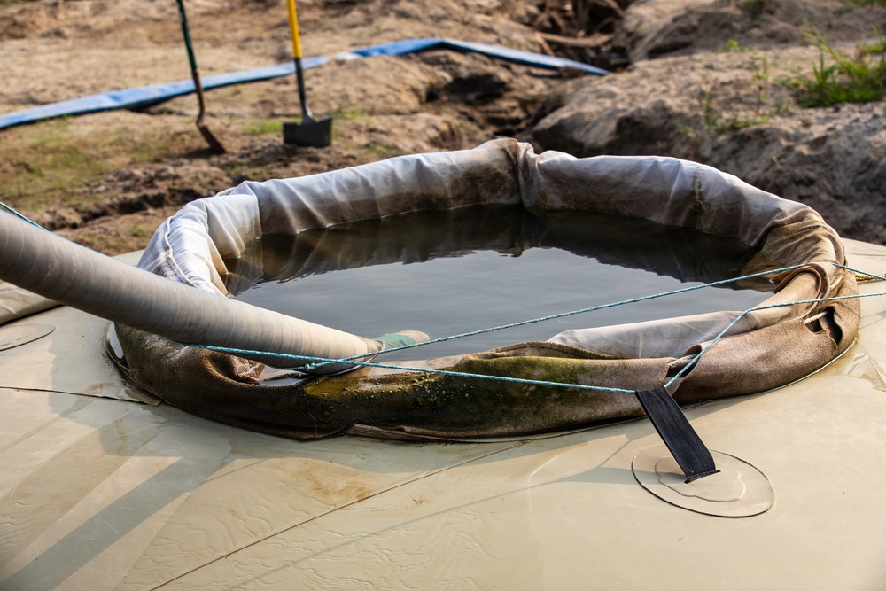 The image shows a circular containment berm or cofferdam filled with dark liquid, likely water or wastewater, situated on top of what appears to be a large tank or bladder. A flexible hose or pipe is inserted into the containment area, suggesting pumping or drainage activity. The setting appears to be an active excavation or construction/remediation site.