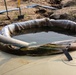 The image shows a circular containment berm or cofferdam filled with dark liquid, likely water or wastewater, situated on top of what appears to be a large tank or bladder. A flexible hose or pipe is inserted into the containment area, suggesting pumping or drainage activity. The setting appears to be an active excavation or construction/remediation site.