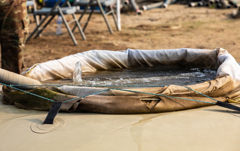 The image shows a military-style collapsible fabric water storage tank or blivet being filled with water via a pipe or hose. Water is actively flowing into the circular, canvas-walled portable reservoir, which is secured with rope or cord ties. The setting appears to be a field operations or training environment with sandy/dirt terrain and equipment visible in the background.