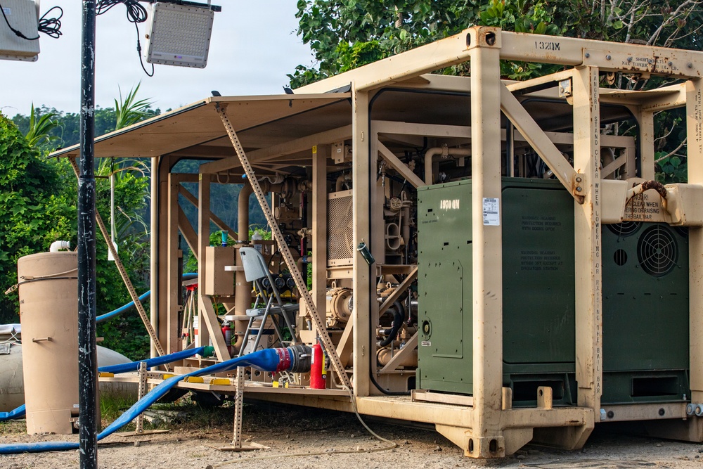 The image shows a military-grade containerized equipment unit, likely a water purification or power generation system, painted in tan/desert camouflage with olive drab green components. The unit is deployed in an outdoor setting with tropical vegetation in the background, suggesting a forward operating base or disaster relief operation. Multiple hoses, pipes, and mechanical components are visible, indicating the system is actively set up for operation.