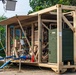 The image shows a military-grade containerized equipment unit, likely a water purification or power generation system, painted in tan/desert camouflage with olive drab green components. The unit is deployed in an outdoor setting with tropical vegetation in the background, suggesting a forward operating base or disaster relief operation. Multiple hoses, pipes, and mechanical components are visible, indicating the system is actively set up for operation.