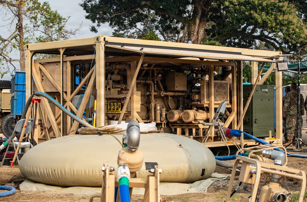 A military field water purification or treatment unit is deployed in an outdoor setting, housed within a tan/sand-colored open-frame containerized shelter. The setup includes pumps, piping, motors, and a collapsible water bladder storage tank in the foreground. At least one uniformed military personnel is visible in the background, suggesting an active field operation.