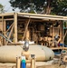 A military field water purification or treatment unit is deployed in an outdoor setting, housed within a tan/sand-colored open-frame containerized shelter. The setup includes pumps, piping, motors, and a collapsible water bladder storage tank in the foreground. At least one uniformed military personnel is visible in the background, suggesting an active field operation.
