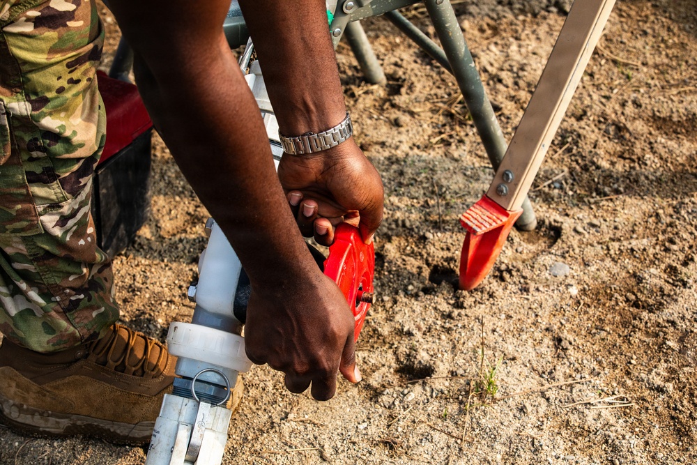 A person wearing military camouflage (OCP/MultiCam pattern) uniform and tan combat boots is working at ground level, manipulating a red valve or fitting on a white PVC pipe assembly. The individual is wearing a silver metal bracelet on their wrist. A metal stand or tripod with a red base plate is visible in the background on sandy/gravelly ground.