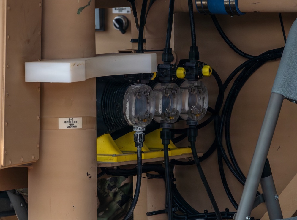 The image shows a close-up of industrial or military equipment featuring fluid handling components mounted on a tan/beige painted metal structure. Three transparent bowl-shaped strainer or filter housings are visible alongside black hoses, yellow fittings, and electrical cables. A label on the structure reads 'S-2 MICROFILTER FEED STRAINER', indicating this is part of a filtration system.