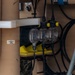 The image shows a close-up of industrial or military equipment featuring fluid handling components mounted on a tan/beige painted metal structure. Three transparent bowl-shaped strainer or filter housings are visible alongside black hoses, yellow fittings, and electrical cables. A label on the structure reads 'S-2 MICROFILTER FEED STRAINER', indicating this is part of a filtration system.