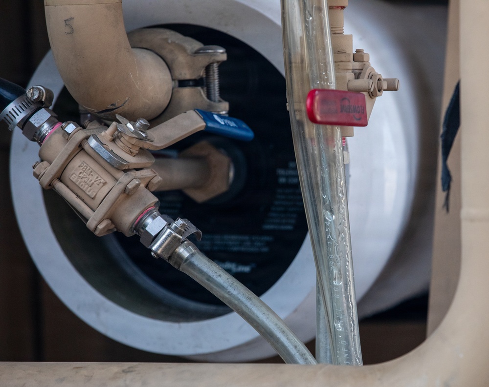 The image shows a close-up of a plumbing assembly, likely associated with a water heater or pressure regulation system. Multiple valves, fittings, and flexible hoses are visible, including a bronze/brass pressure reducing valve and a blue-handled ball valve. The setup appears to be a residential or light commercial water supply connection point.