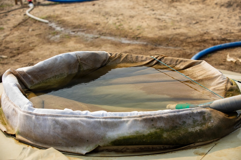 The image shows a portable, collapsible fabric water storage tank or bladder partially filled with murky water, situated on dry, sandy ground. A blue hose and a green fitting are visible connecting to the tank, suggesting active water supply or transfer operations. The tank appears weathered and heavily used, with visible dirt, algae, and wear on its fabric walls.