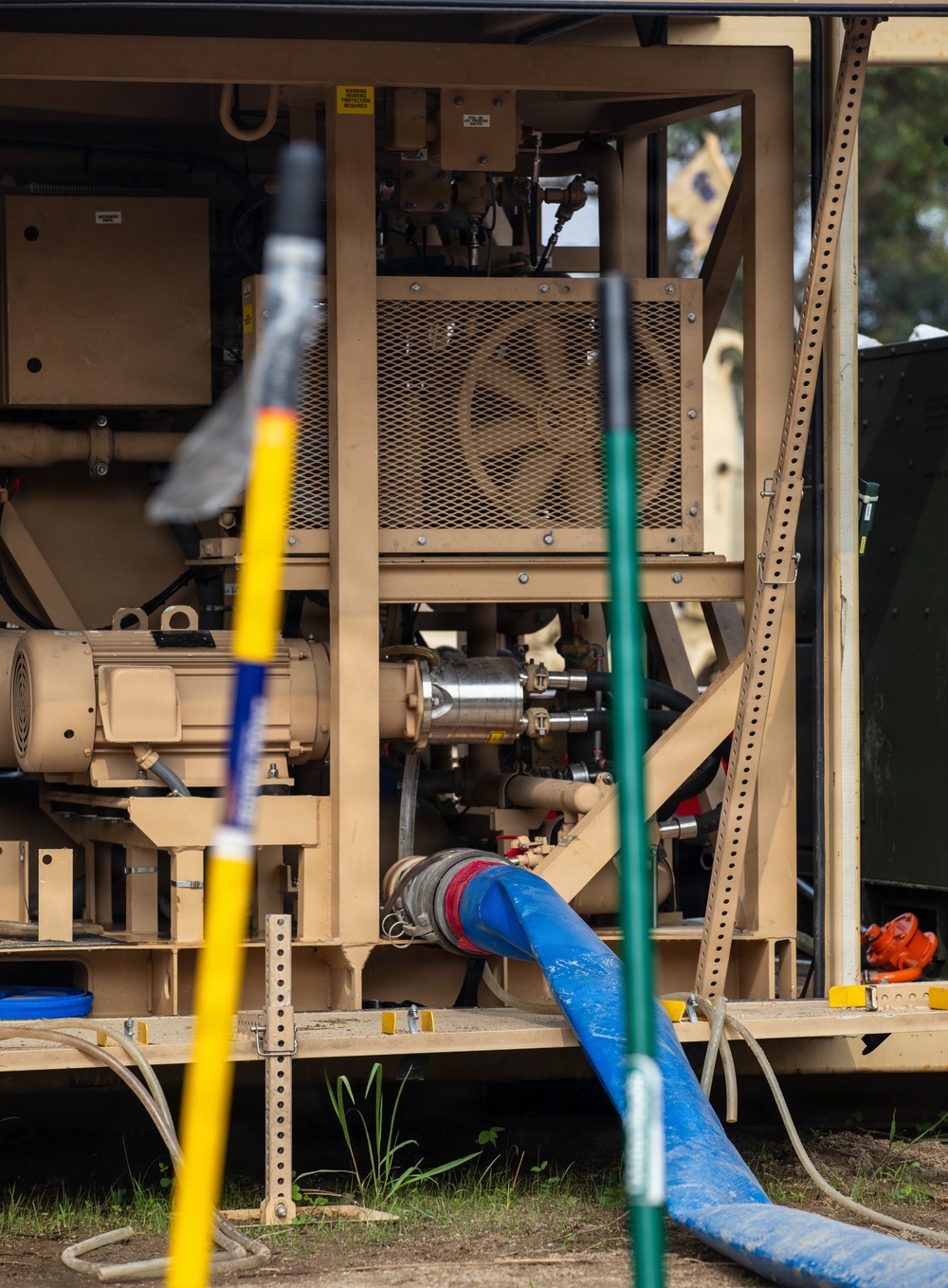 The image shows a tan/beige military or industrial pump unit mounted on a skid frame, with various hoses and mechanical components visible. A large blue flexible hose is connected to the pump assembly, and two colored poles (yellow and green) are positioned in the foreground. The equipment appears to be deployed outdoors on a grassy/dirt surface.
