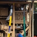The image shows a tan/beige military or industrial pump unit mounted on a skid frame, with various hoses and mechanical components visible. A large blue flexible hose is connected to the pump assembly, and two colored poles (yellow and green) are positioned in the foreground. The equipment appears to be deployed outdoors on a grassy/dirt surface.