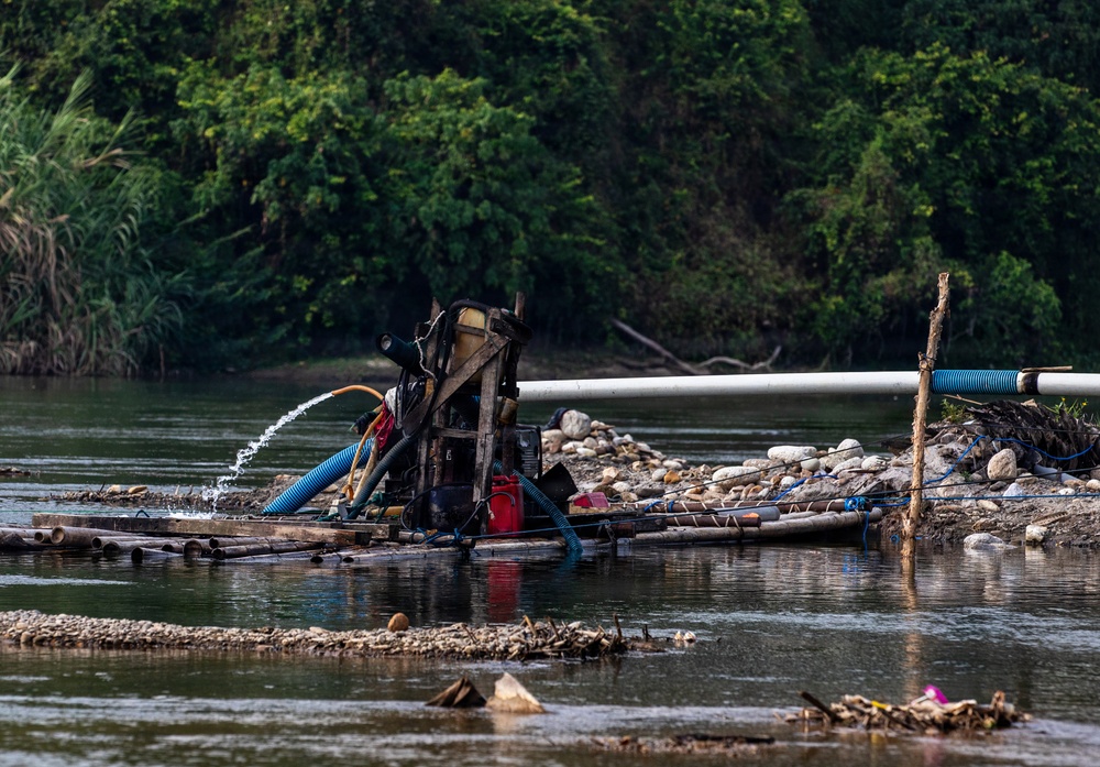A makeshift river dredging or mining operation is visible on a shallow river, featuring a mechanical pump mounted on a bamboo raft. The equipment is actively pumping water, with hoses and pipes extending across a gravel and rock bar. The setting appears to be a tropical or subtropical river environment with dense green vegetation in the background.
