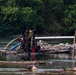 A makeshift river dredging or mining operation is visible on a shallow river, featuring a mechanical pump mounted on a bamboo raft. The equipment is actively pumping water, with hoses and pipes extending across a gravel and rock bar. The setting appears to be a tropical or subtropical river environment with dense green vegetation in the background.