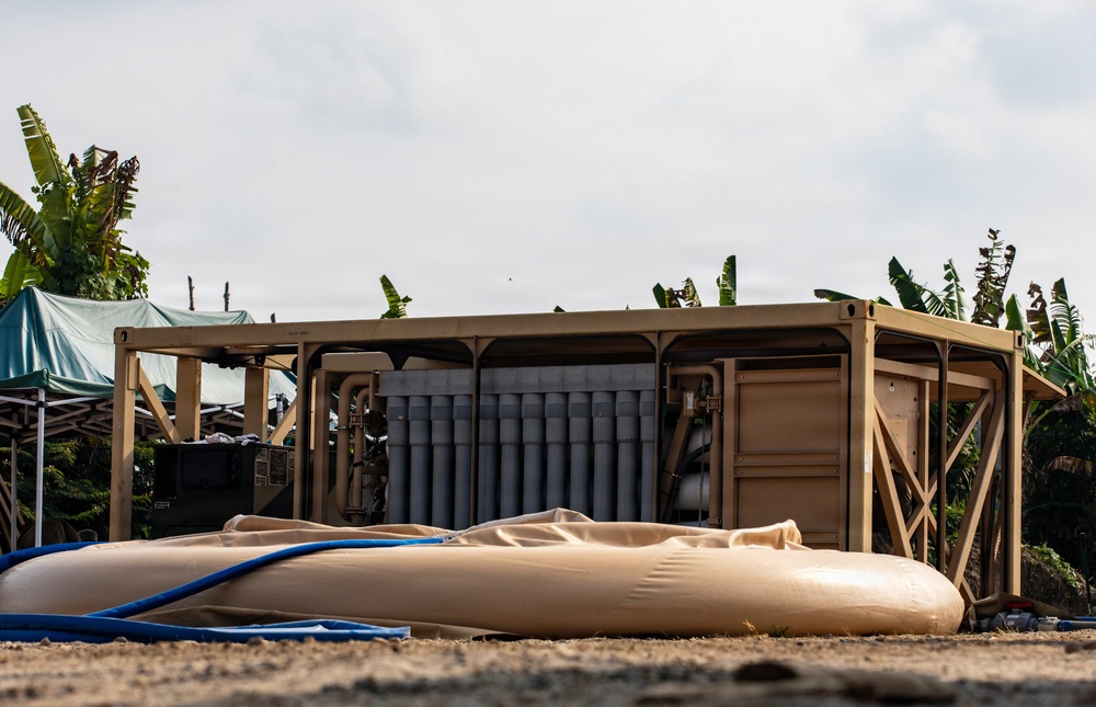 The image shows a military or humanitarian field installation featuring a tan/sand-colored open-frame equipment shelter containing cylindrical filter or storage units and piping. In the foreground, deflated or partially inflated tan flexible bladder tanks are visible on sandy ground, connected by blue hoses. The setting appears to be a tropical location, indicated by banana trees in the background, suggesting a disaster relief or expeditionary water purification operation.