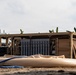 The image shows a military or humanitarian field installation featuring a tan/sand-colored open-frame equipment shelter containing cylindrical filter or storage units and piping. In the foreground, deflated or partially inflated tan flexible bladder tanks are visible on sandy ground, connected by blue hoses. The setting appears to be a tropical location, indicated by banana trees in the background, suggesting a disaster relief or expeditionary water purification operation.