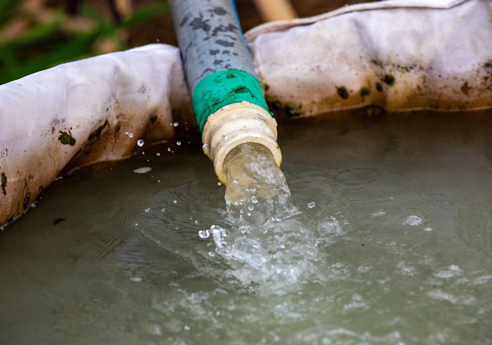The image shows water actively flowing from a pipe or hose fitting into a contained water reservoir or tank. The pipe has a green coupling connector joining two sections, and the surrounding fabric appears to be part of a portable water storage bladder or containment bag. The water appears slightly turbid or murky, suggesting it may be groundwater or irrigation water.