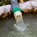 The image shows water actively flowing from a pipe or hose fitting into a contained water reservoir or tank. The pipe has a green coupling connector joining two sections, and the surrounding fabric appears to be part of a portable water storage bladder or containment bag. The water appears slightly turbid or murky, suggesting it may be groundwater or irrigation water.