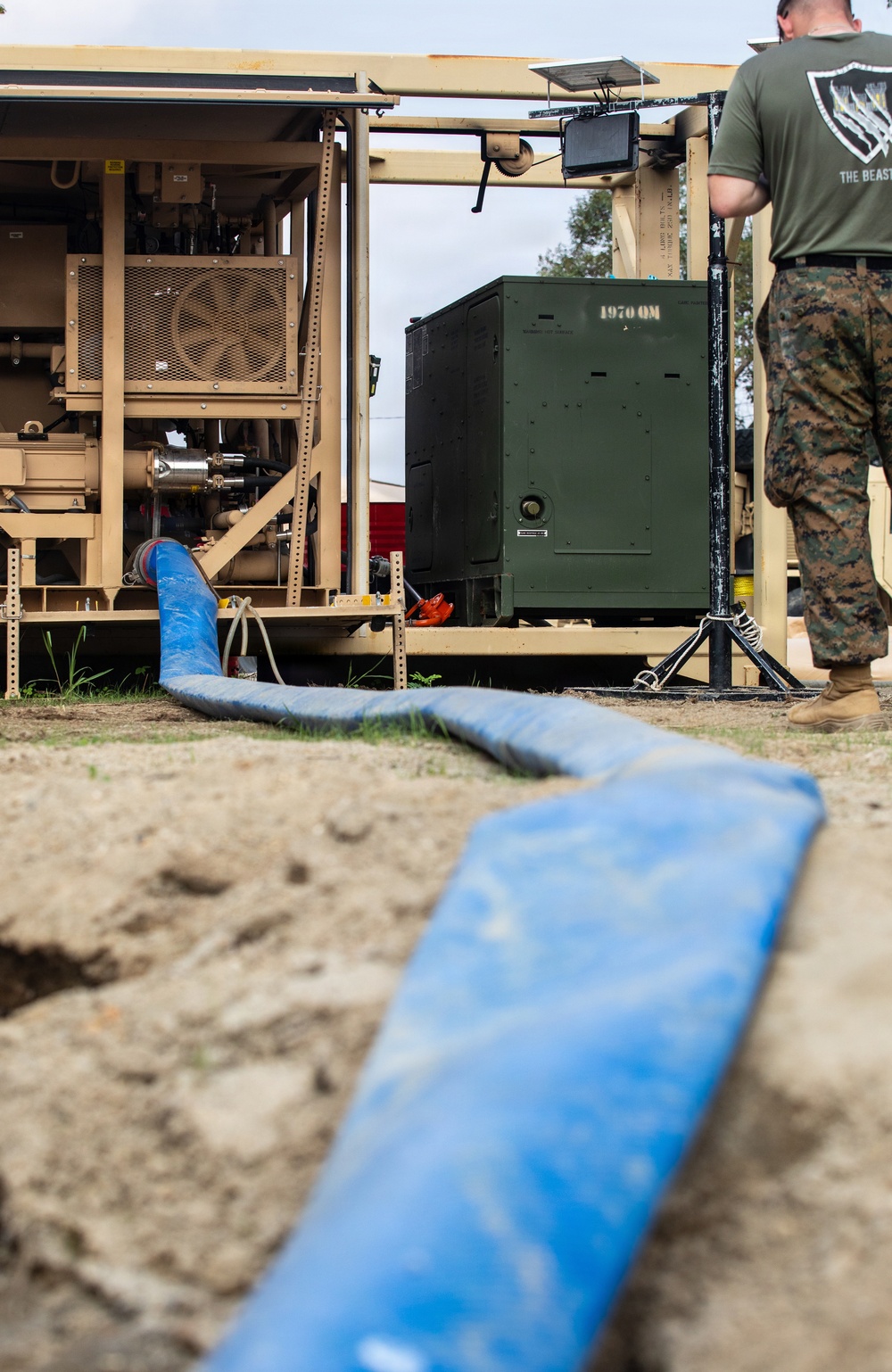 A military service member in Marine Corps camouflage utilities and a green 'THE BEAST' t-shirt stands beside tan-colored military equipment, likely a water purification or pumping system. A large blue flexible hose runs from the equipment across sandy ground in the foreground. A dark olive-green military container marked '1970 QM' is visible within the equipment frame.