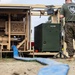 A military service member in Marine Corps camouflage utilities and a green 'THE BEAST' t-shirt stands beside tan-colored military equipment, likely a water purification or pumping system. A large blue flexible hose runs from the equipment across sandy ground in the foreground. A dark olive-green military container marked '1970 QM' is visible within the equipment frame.