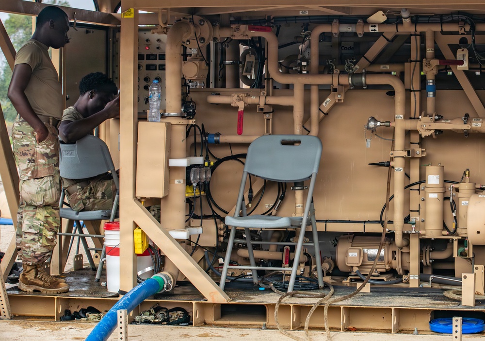 Two military personnel in multicam/OCP uniforms are working inside what appears to be a military water purification or fluid management unit, painted in tan/desert color. The unit contains an extensive network of pipes, valves, pumps, and control panels. The setting appears to be an outdoor forward operating base or field environment.