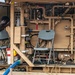 Two military personnel in multicam/OCP uniforms are working inside what appears to be a military water purification or fluid management unit, painted in tan/desert color. The unit contains an extensive network of pipes, valves, pumps, and control panels. The setting appears to be an outdoor forward operating base or field environment.