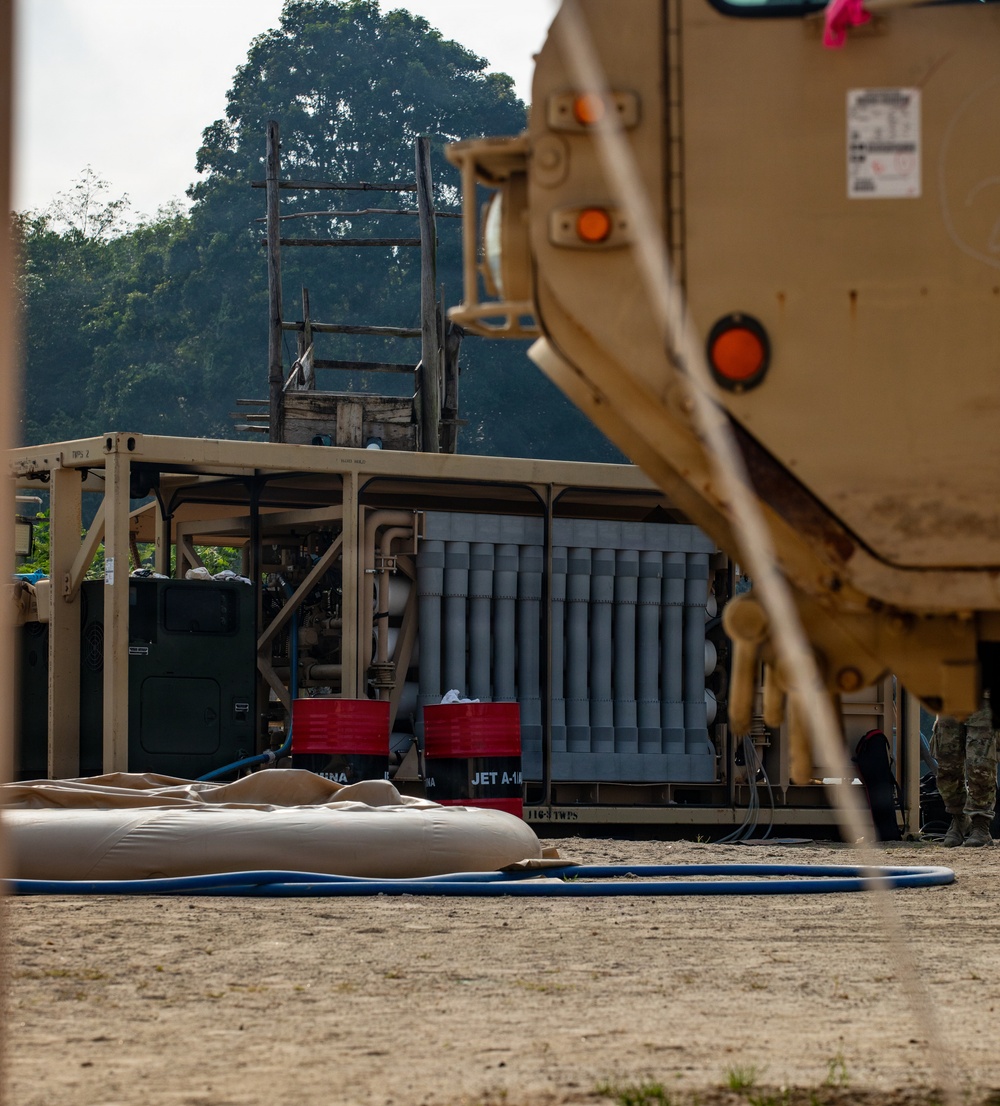 The image depicts a military forward operating base or fuel point, featuring tactical petroleum handling equipment including what appears to be a Tactical Water Purification System (TWPS) or fuel distribution unit with visible 'JET A-1' labeling. A tan military vehicle occupies the right foreground, partially obscuring the scene. Collapsible fuel or water bladders and blue hoses are laid out on the ground, indicating active logistics or refueling operations.