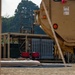 The image depicts a military forward operating base or fuel point, featuring tactical petroleum handling equipment including what appears to be a Tactical Water Purification System (TWPS) or fuel distribution unit with visible 'JET A-1' labeling. A tan military vehicle occupies the right foreground, partially obscuring the scene. Collapsible fuel or water bladders and blue hoses are laid out on the ground, indicating active logistics or refueling operations.